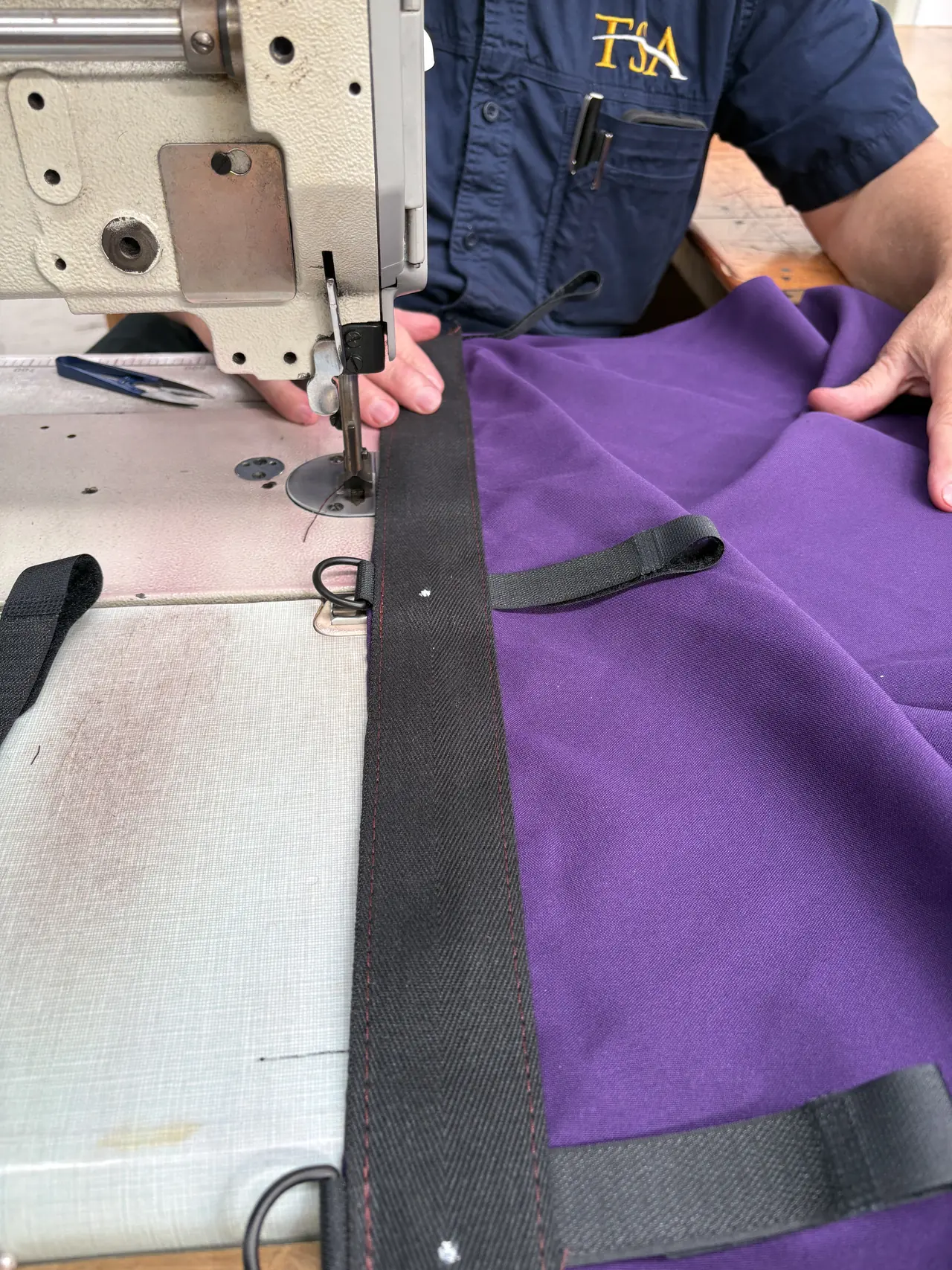 Black webbing with a Velcro tab and a D-ring being sewn onto the edge of a purple roof swag at an industrial sewing machine